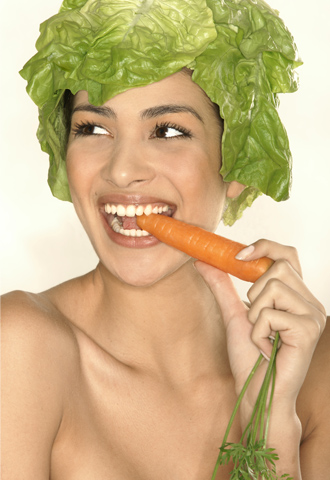 Dieses Bild steht für die biologische Zahnmedizin. Es zeigt eine Frau. Ihr Kopf ist von grünen Salatblättern umhüllt. Ihre Zähne beißen auf ein frische Möhre. 
